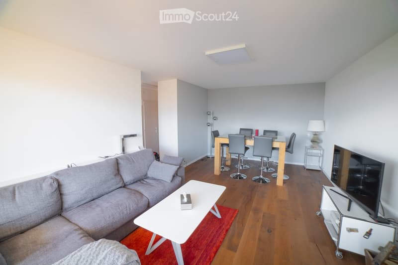 Open-plan living and dining area with a grey sectional sofa, white coffee table, dining table with chairs, and a TV stand with a flat-screen TV.