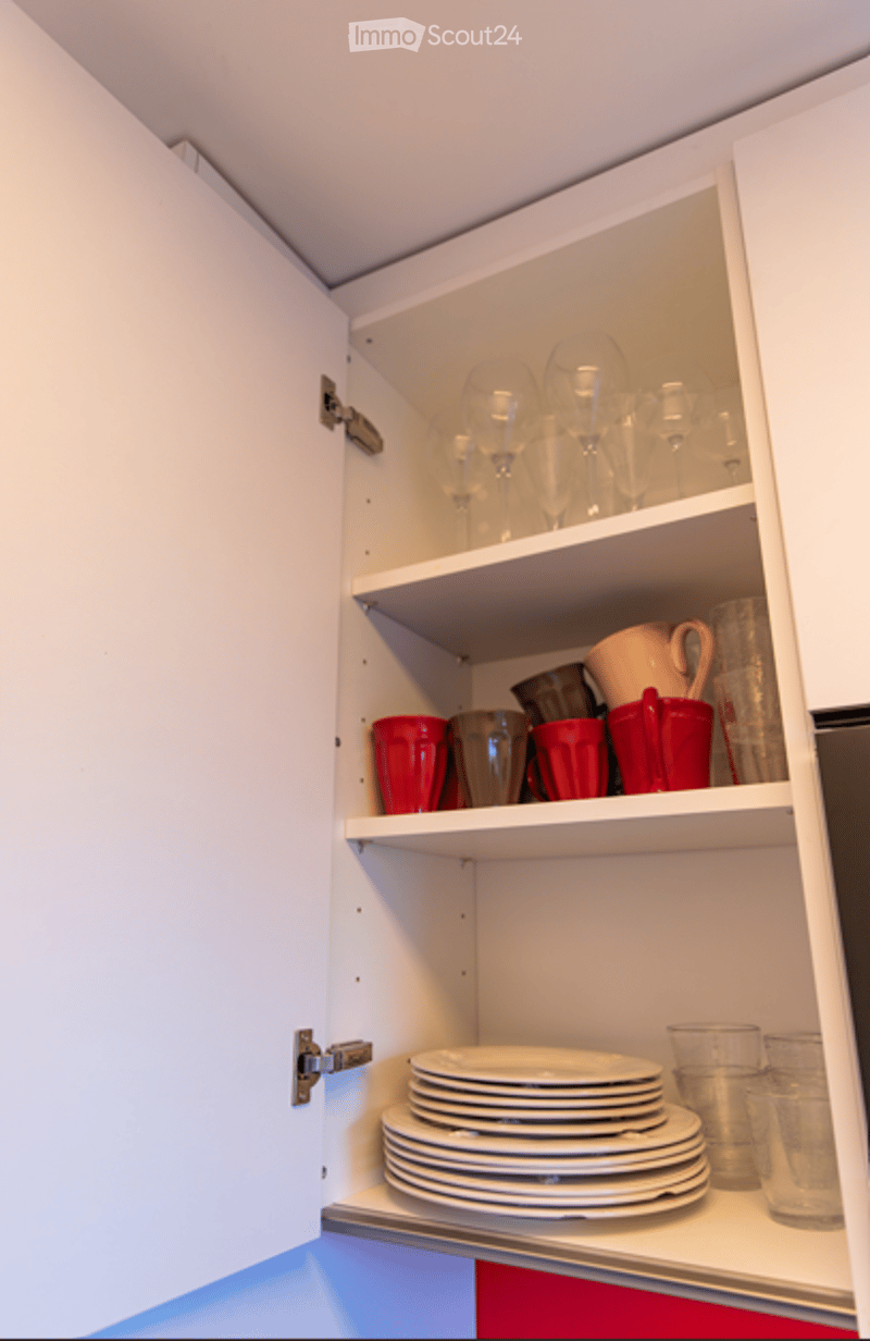 Open kitchen cabinet displaying neatly stacked plates, glasses, mugs, and cups on multiple shelves.
