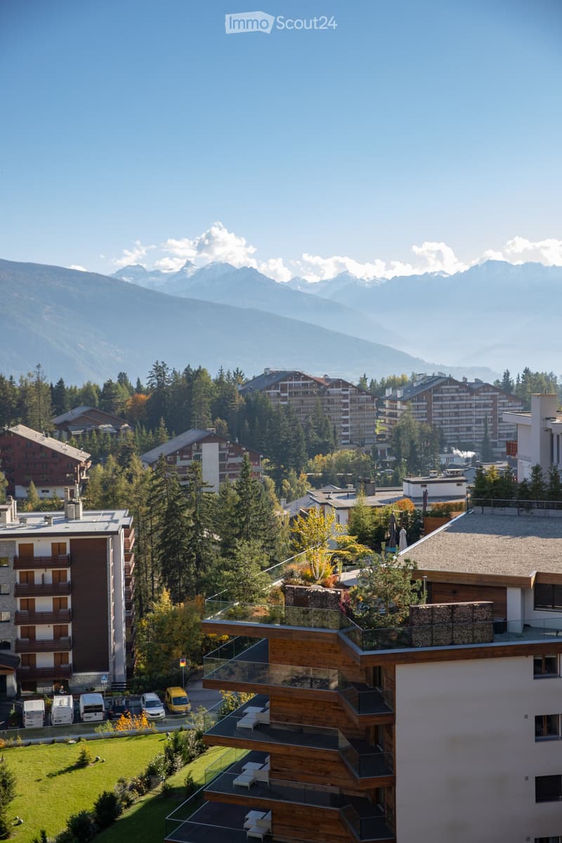 A modern apartment building with multiple balconies and a rooftop terrace, surrounded by trees and other buildings, with a scenic mountain view in the background.