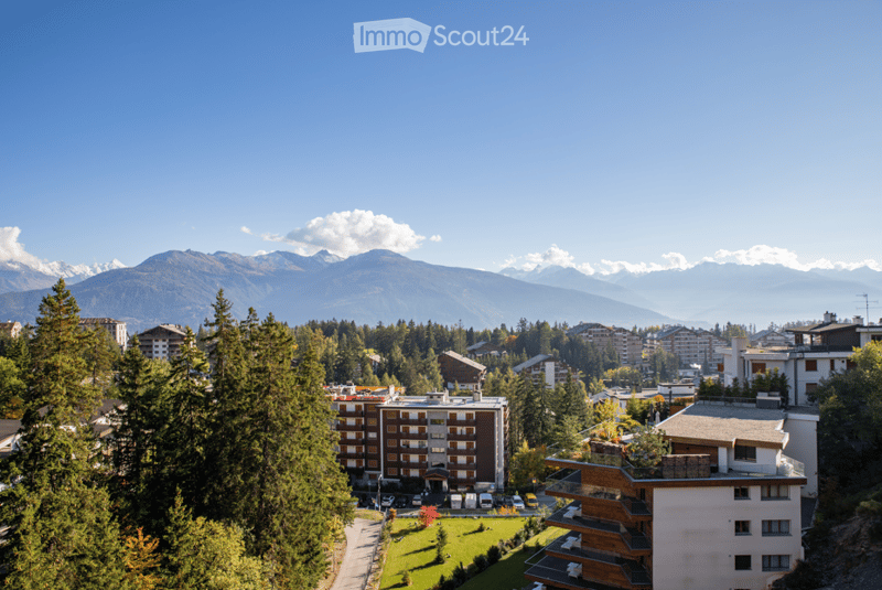 A scenic view of a town nestled in a valley with snow-capped mountains in the background, surrounded by dense forests and modern residential buildings.