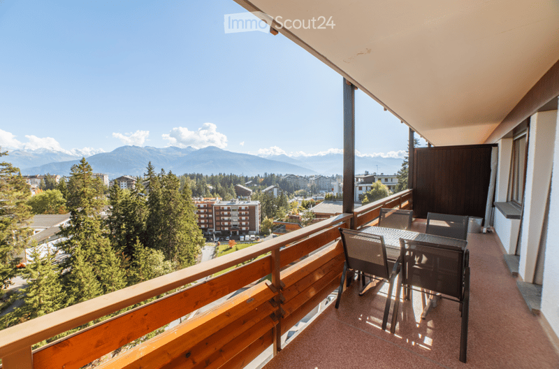 Balcony with wooden railing, overlooking a scenic mountain view and town below, furnished with a table and chairs.