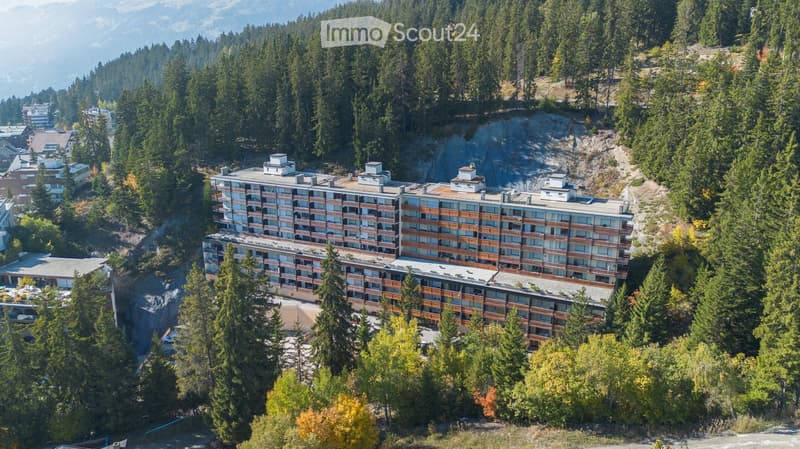 A multi-story hotel or apartment complex surrounded by dense forest and mountains, with balconies and a parking area.