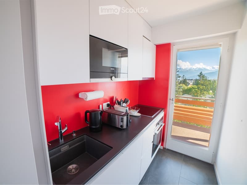 Modern kitchen with white cabinets, red accent wall, black countertop, and a view of mountains through a large window. Includes a sink, oven, microwave, toaster, and various small appliances.