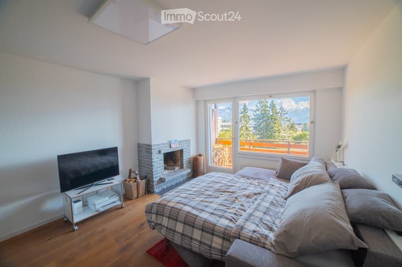 Cozy bedroom with a large bed, plaid blanket, pillows, a television on a stand, a brick fireplace, and a scenic mountain view through large windows.