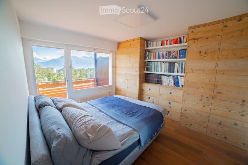 Bright bedroom with wooden flooring, large window offering mountain view, bed with pillows and blanket, built-in wooden bookshelf, and drawers.