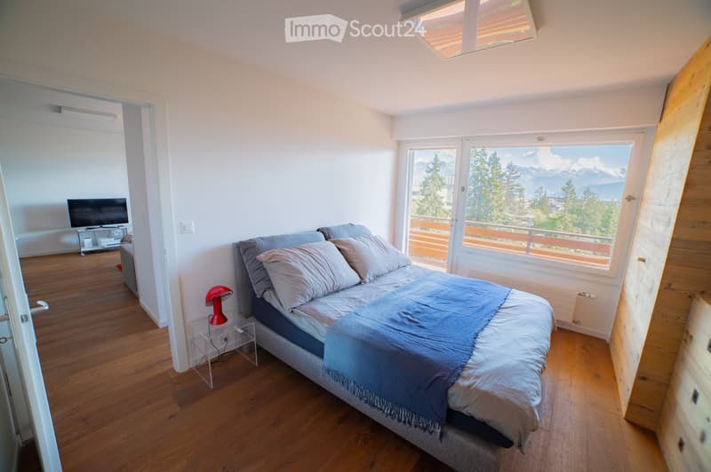 Bright bedroom with wooden flooring, large window offering a scenic mountain view, bed with pillows and blanket, table lamp, nightstand, television, and an open doorway leading to another room.