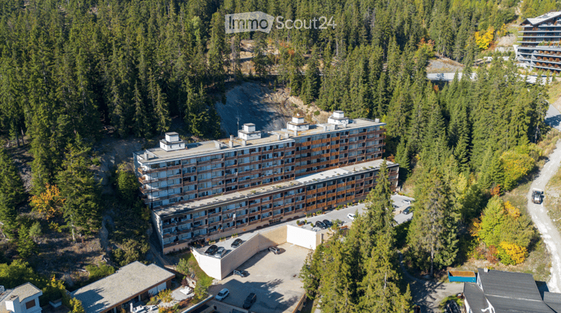 Aerial view of a multi-story residential building surrounded by dense forest, with a parking area and several cars visible.