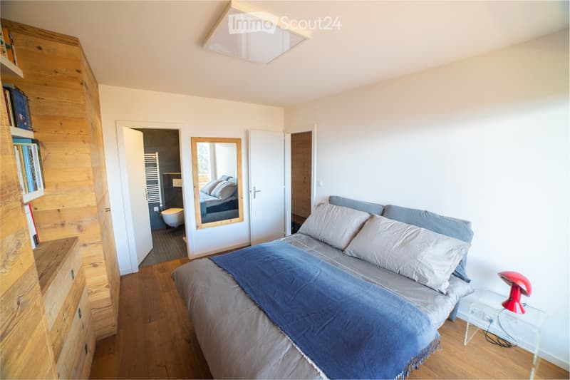 Cozy bedroom with a wooden bed, nightstand, red lamp, and a large mirror. A wooden wardrobe with books is visible, and an adjacent bathroom is partially visible through an open door.