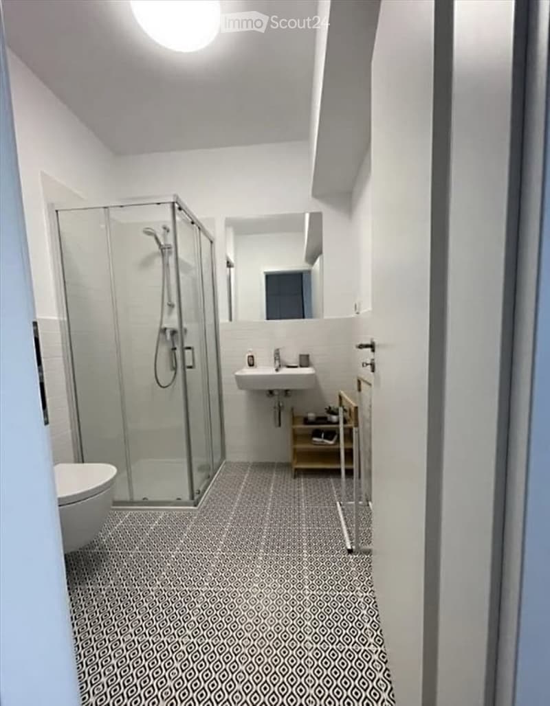 Modern bathroom with white walls, grey patterned tile floor, glass-enclosed shower, toilet, sink, and a small wooden shelf.