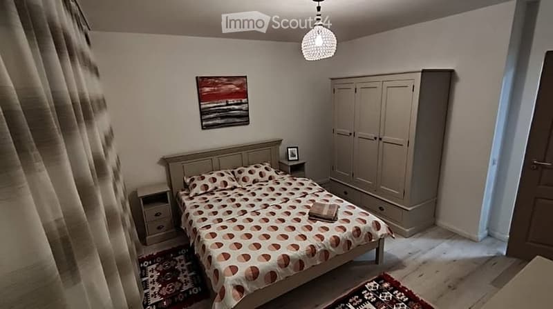 Cozy bedroom with a double bed, nightstand, wardrobe, and a decorative painting on the wall. The room features wooden flooring and a modern chandelier.