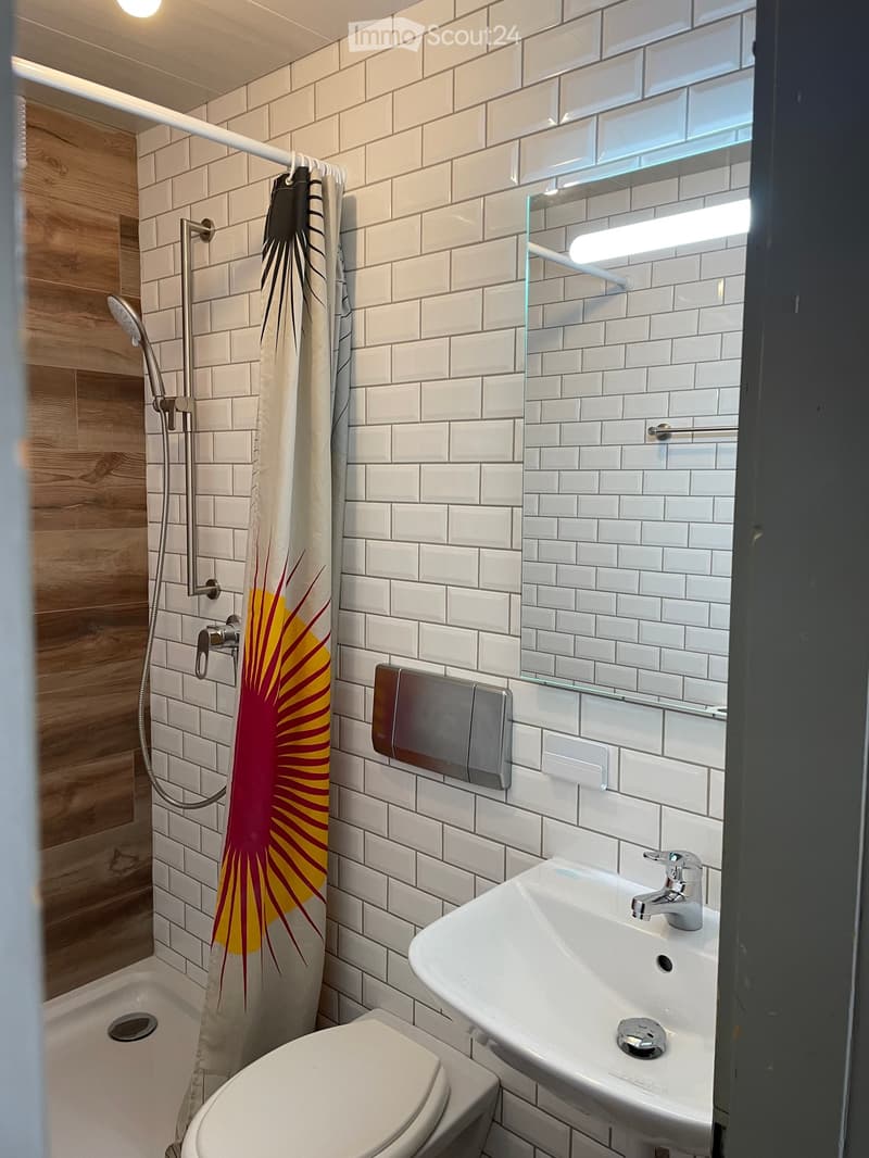 Modern bathroom with white tiled walls, a shower with a decorative curtain, a toilet, a sink with a modern faucet, and a built-in light fixture above the mirror.