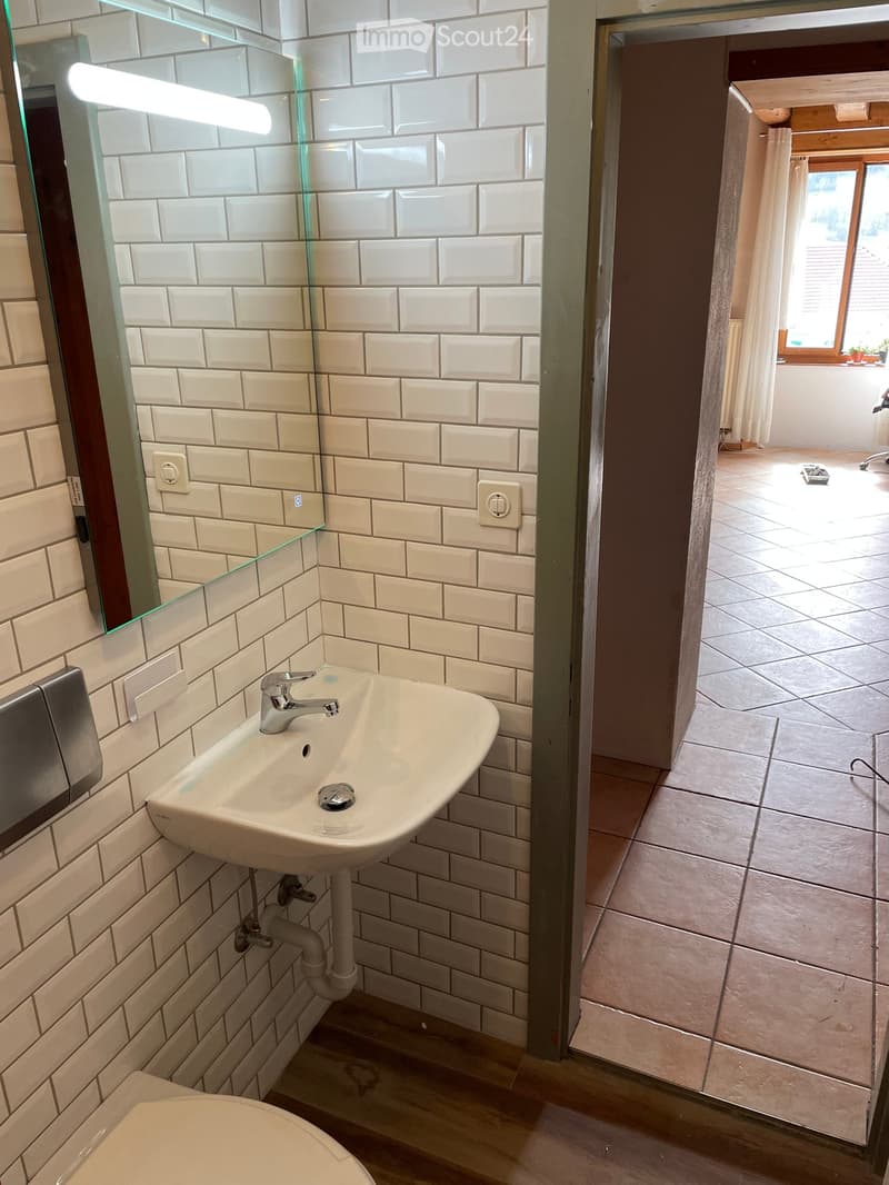 Modern bathroom with white tiled walls, a white sink with a chrome faucet, a mirror, and a toilet. The floor is tiled, and the room has a wooden door frame leading to another space.