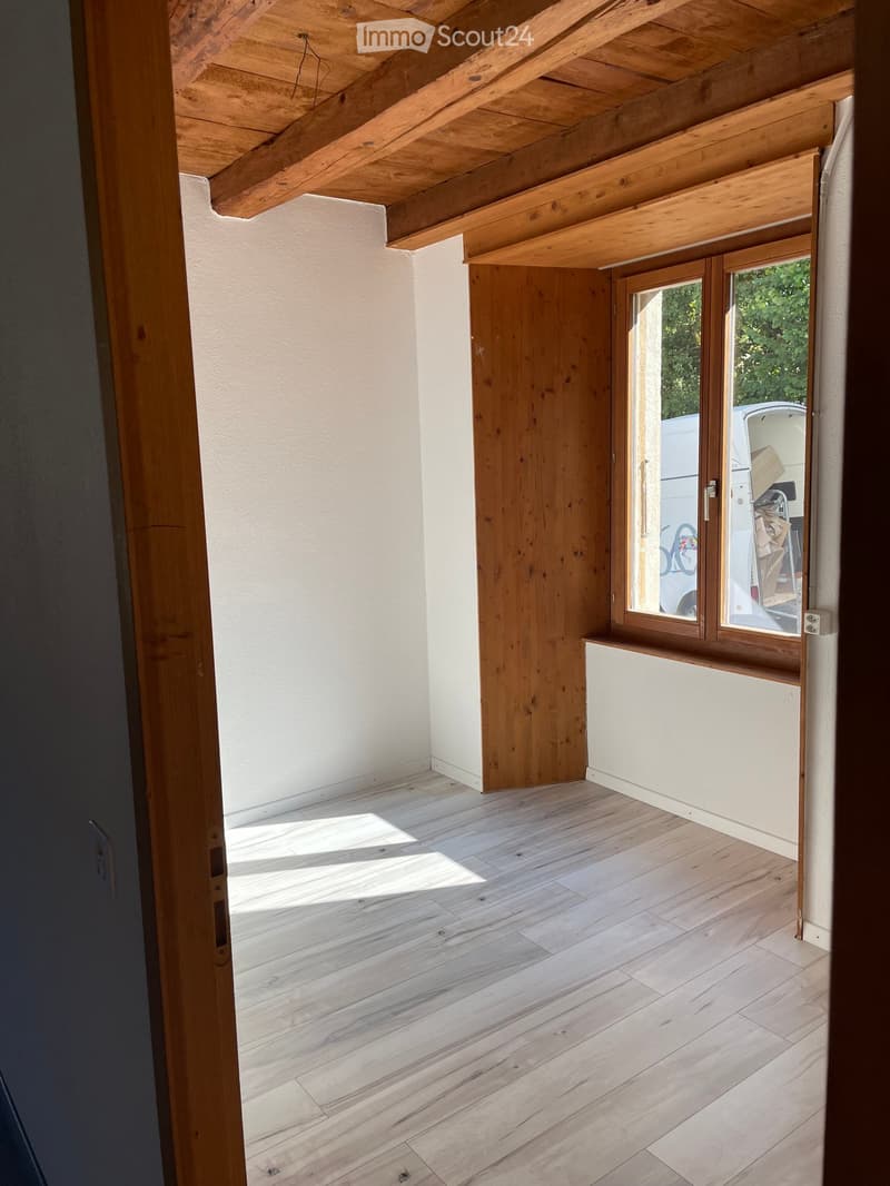 Empty room with wooden floor, white walls, exposed wooden ceiling beams, and a large window with wooden frame leading to an outdoor area.
