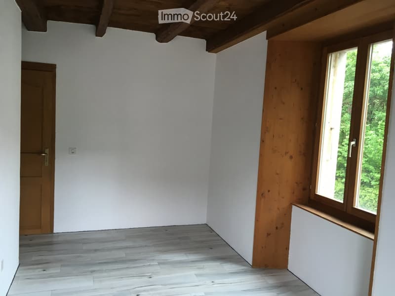 Empty room with light wooden flooring, white walls, and large windows with wooden frames. Features a wooden door and exposed wooden ceiling beams.