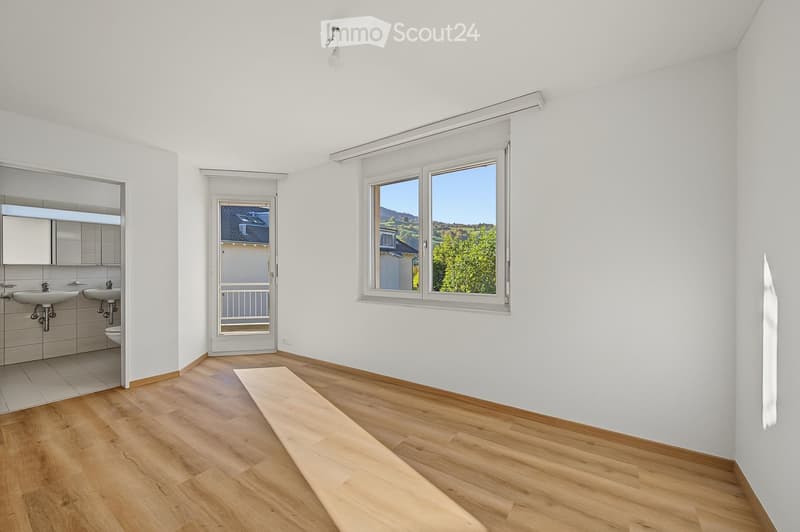Empty room with wooden flooring, white walls, large windows, and a door leading to a balcony. The adjacent bathroom area features two sinks, a toilet, and a mirror.