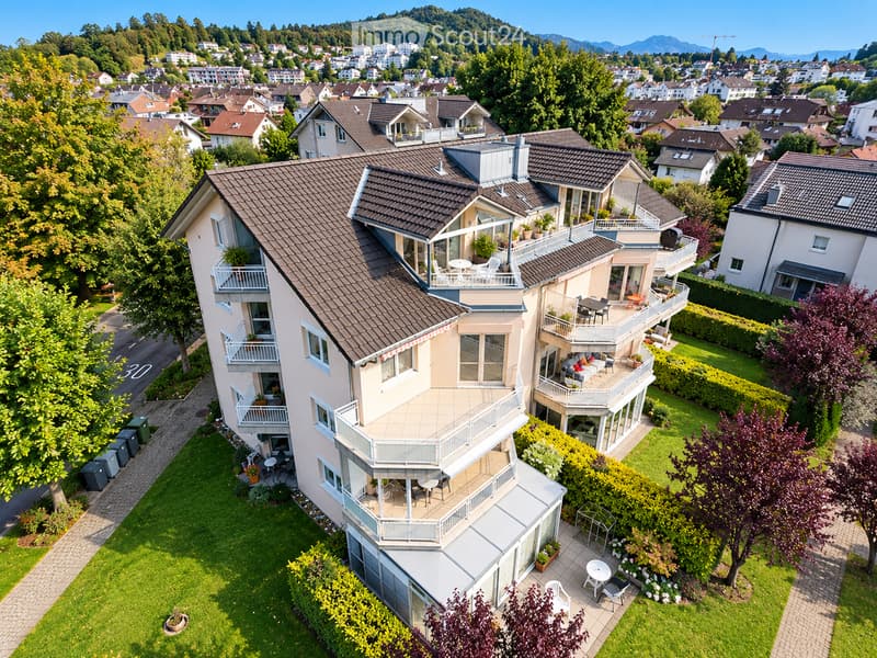 A modern, multi-story house with a brown tiled roof, multiple balconies, and a well-maintained garden. The house features large windows and is surrounded by greenery and neighboring houses in a suburban setting.