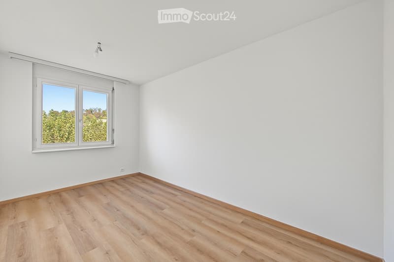 Empty room with white walls, wooden flooring, and a large window offering a view of greenery outside.