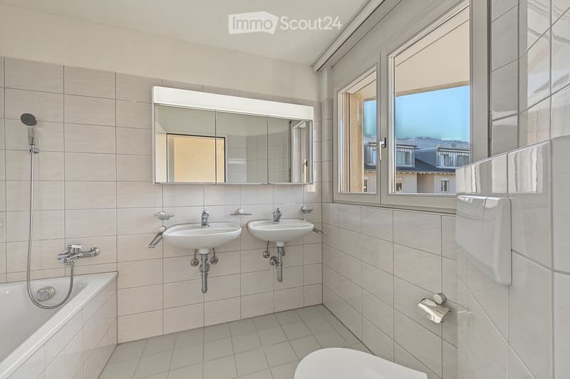Modern bathroom with beige tiles, a bathtub, two sinks, a large mirror, and a toilet. Large windows offer a view of the outside.