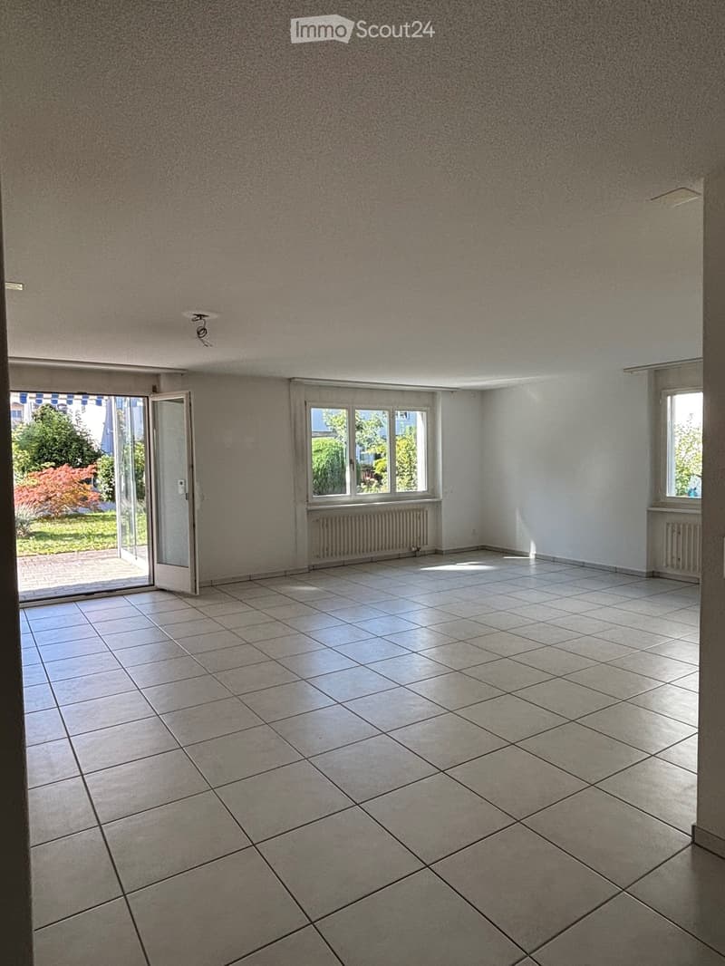 Empty room with tiled floor, large windows, and a sliding glass door leading to a garden area.