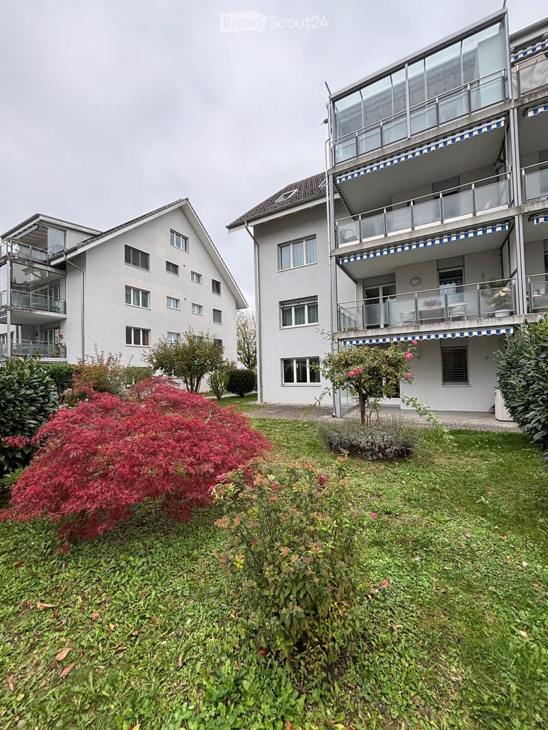 Two-story residential building with balconies, surrounded by a well-maintained garden with shrubs and trees.
