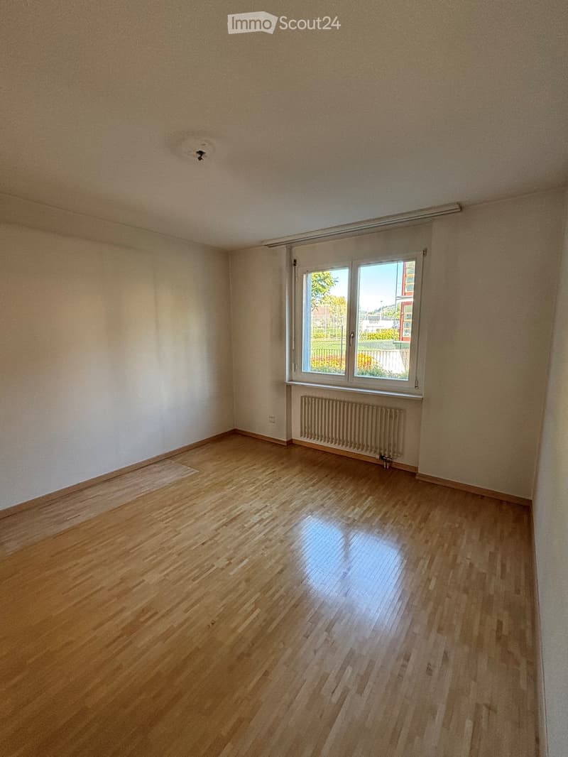 Empty room with wooden flooring, white walls, and a large window with a radiator below it.