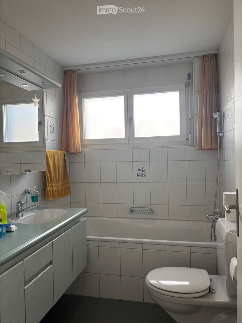 Modern bathroom with a sink, bathtub, toilet, and mirror. Features tiled walls, a window with curtains, and a towel hanging on the wall.