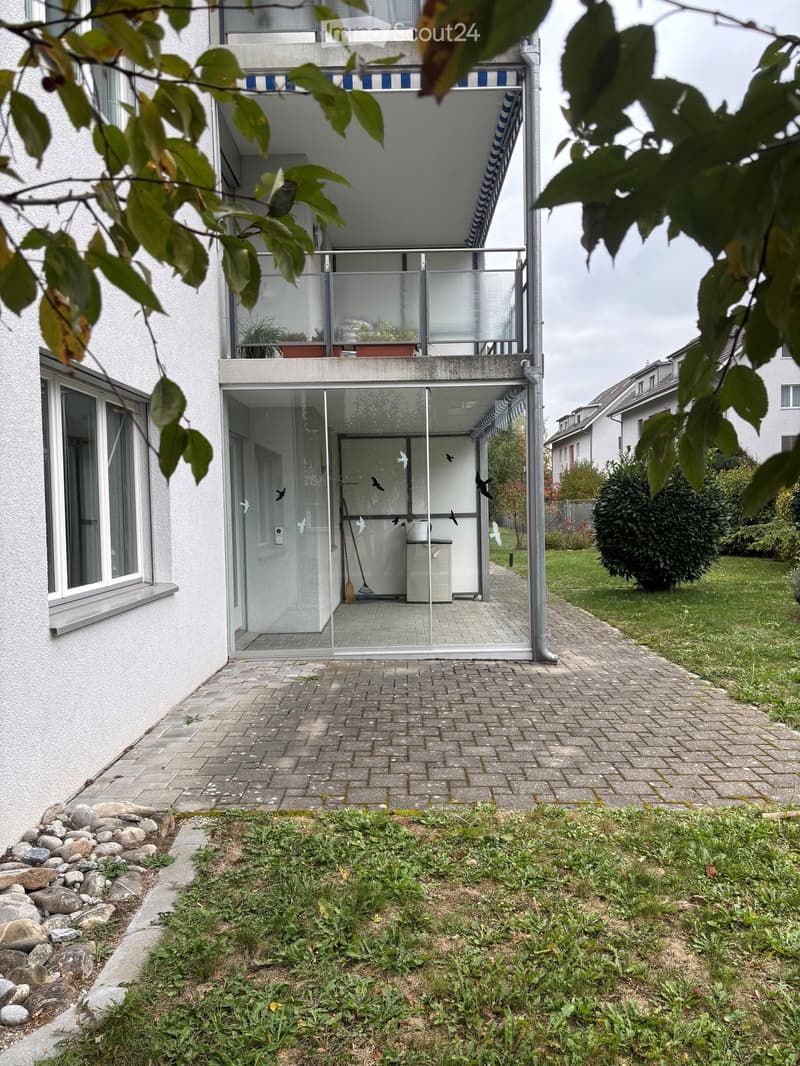 Balcony with glass railing, small patio area with broom and dustpan, adjacent grassy area with rocks and plants.
