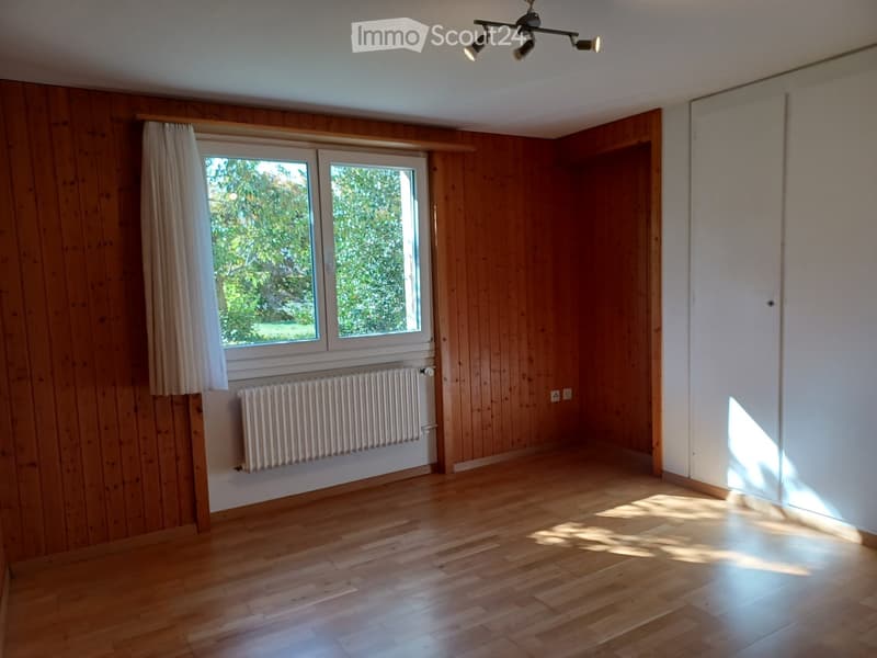 Empty room with wooden walls, wooden floor, large window with white curtain, radiator, and ceiling light fixture.