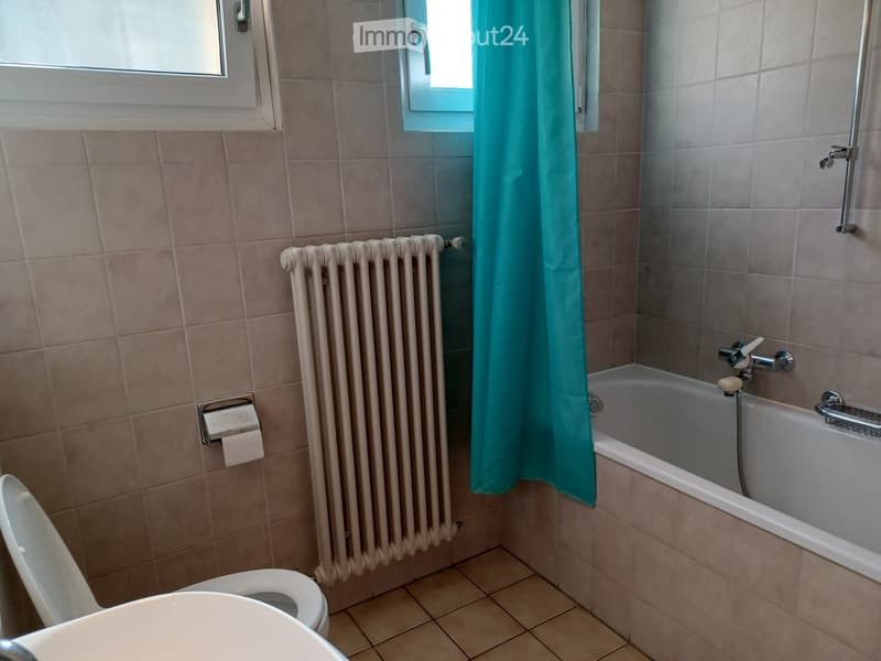 Bathroom with beige tiled walls and floor, featuring a toilet, bathtub with a shower curtain, a radiator, and a toilet paper holder.