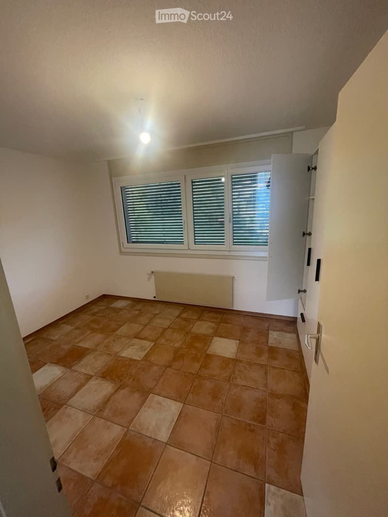 Empty room with tiled floor, large window with blinds, radiator, and open door leading to another space.