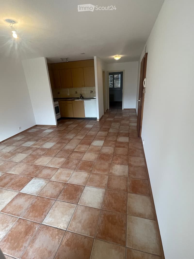Empty hallway with tiled floor, beige walls, and a small kitchen area visible at one end. The kitchen has light-colored cabinets, a sink, and a refrigerator.