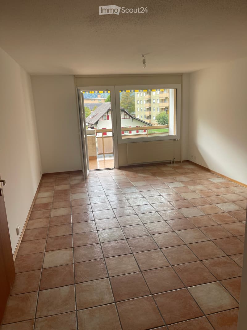 Empty room with tiled floor, white walls, and an open door leading to a balcony with a view of buildings and trees.