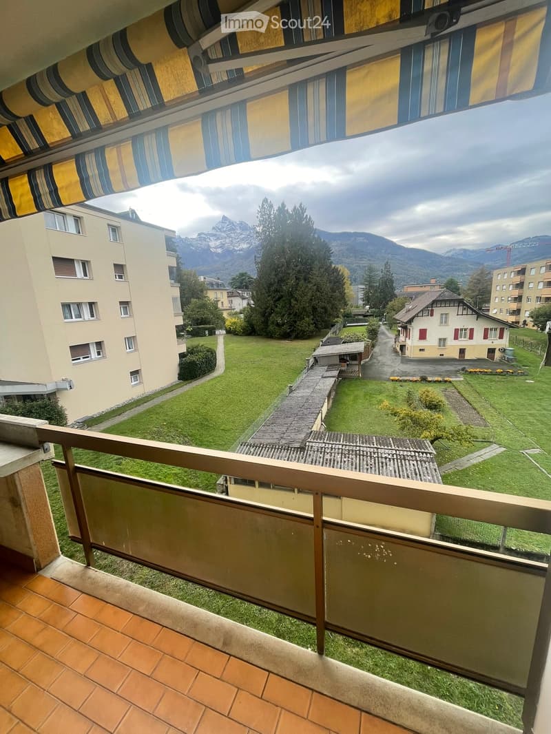Balcony with wooden railing, tiled floor, and a view of a green lawn, trees, and mountains in the background.