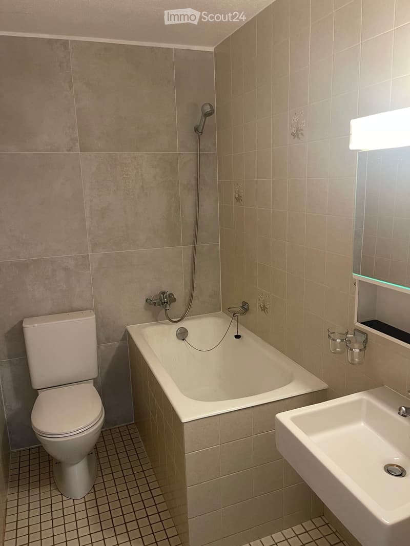 Modern bathroom with beige tiled walls, a bathtub, toilet, sink, and a mirror with shelves holding cups.