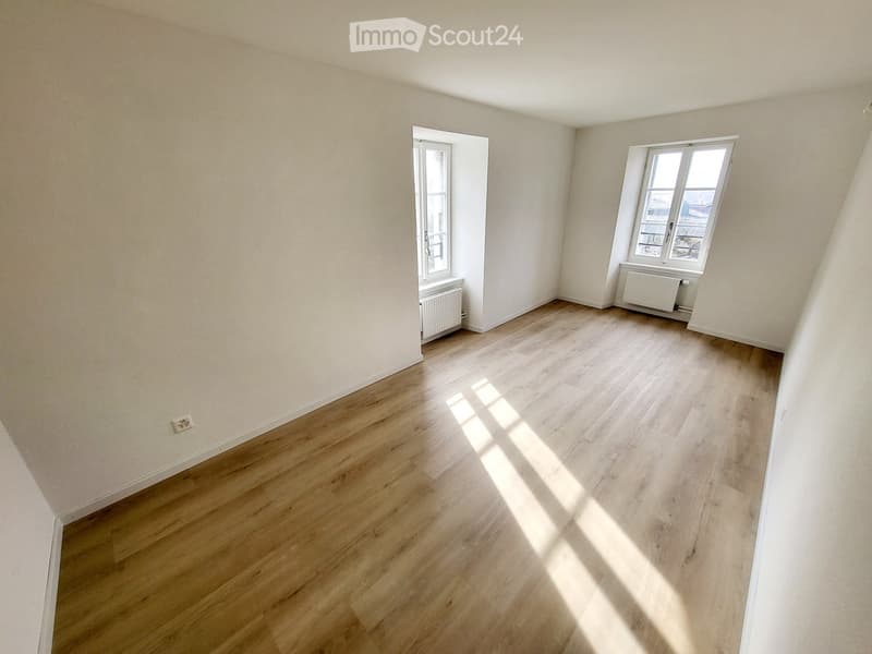 Empty room with light wooden flooring and white walls, featuring two windows and radiators.