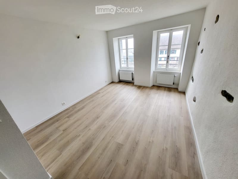 Empty room with light wooden flooring, white walls, and two windows with radiators beneath them.