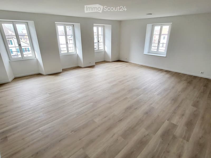 Empty room with light wooden flooring and white walls, featuring multiple windows with white frames and a visible view of neighboring buildings.
