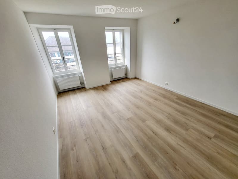 Empty room with light wooden flooring, white walls, and two windows with radiators beneath them.