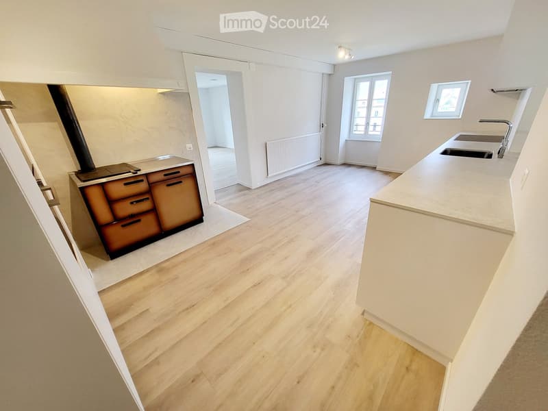 Empty kitchen with wooden flooring, white walls, and a modern design. Features include a built-in stove, a sink, and a window with a view.