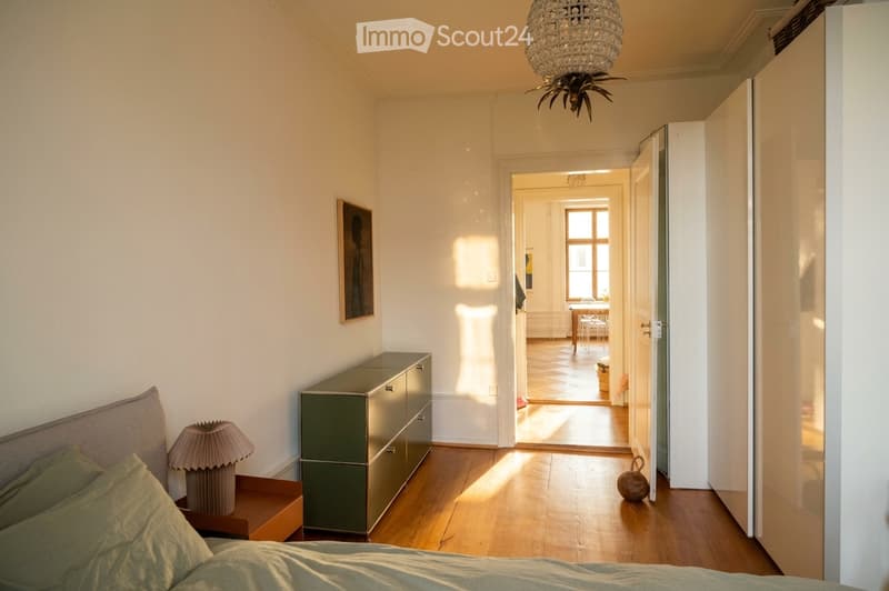 Bright bedroom with wooden flooring, a bed with green bedding, a nightstand with a lamp, a green drawer unit, and an open doorway leading to another room with a table and chairs.