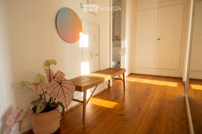 A well-lit hallway with wooden flooring, a small bench, a potted plant, and a colorful wall mirror. The hallway leads to a bathroom with white tiles and a toilet visible in the background.