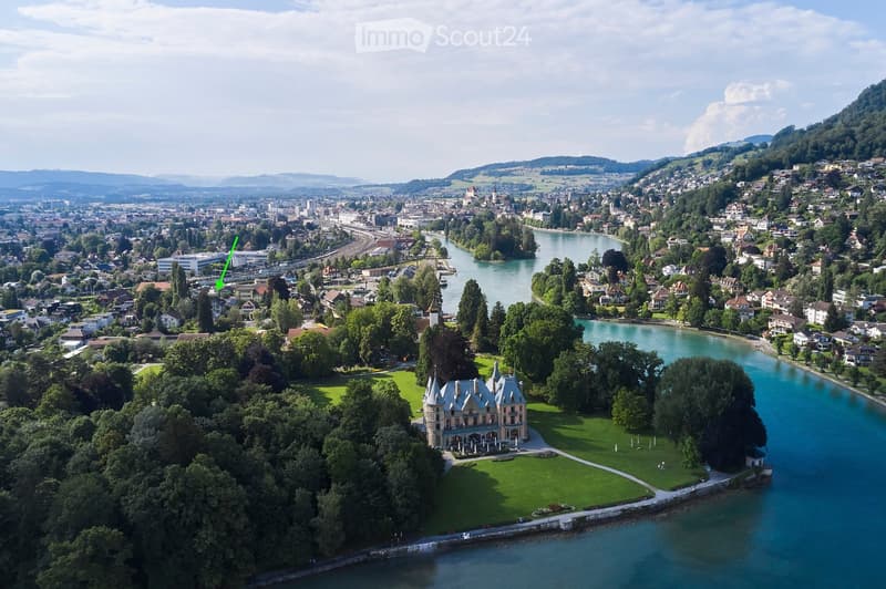 Aerial view of a city with a river running through it, surrounded by green hills and scattered buildings. A prominent mansion with a tower is situated on a grassy area near the riverbank.