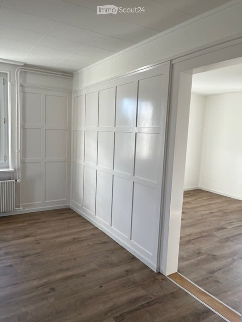 Empty room, wooden floors, white walls, white paneling, door, window, and radiator