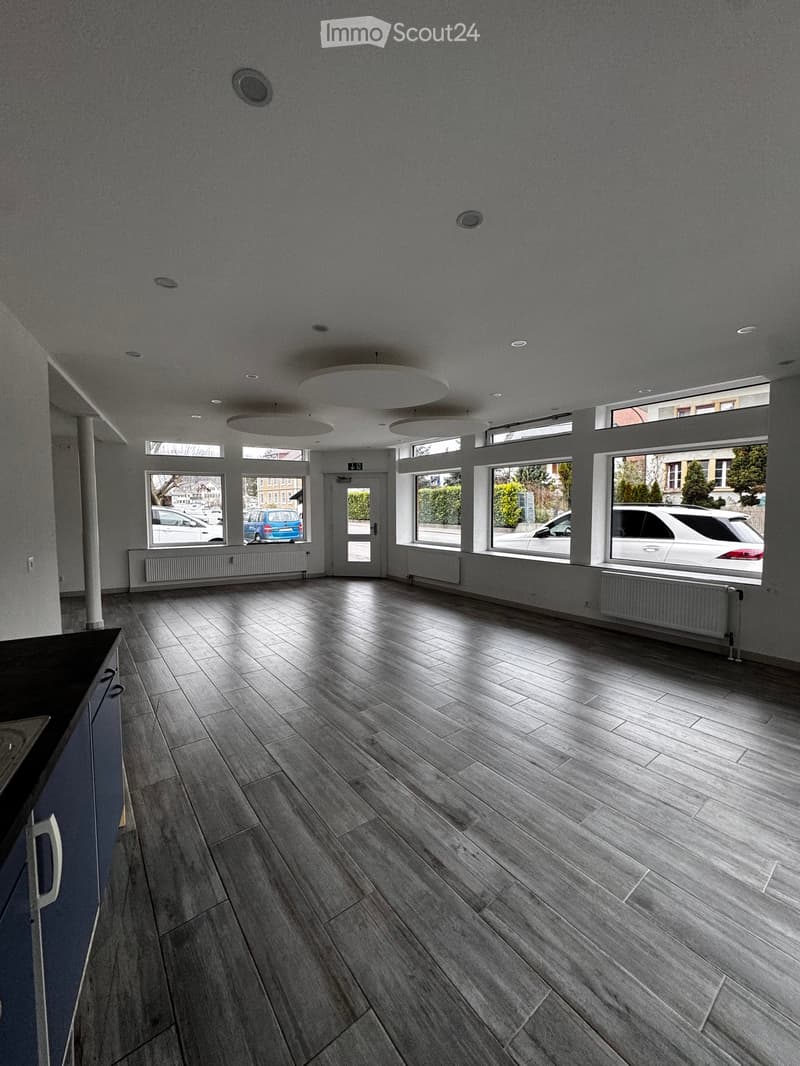 Empty room with wooden flooring, large windows, and modern lighting fixtures.