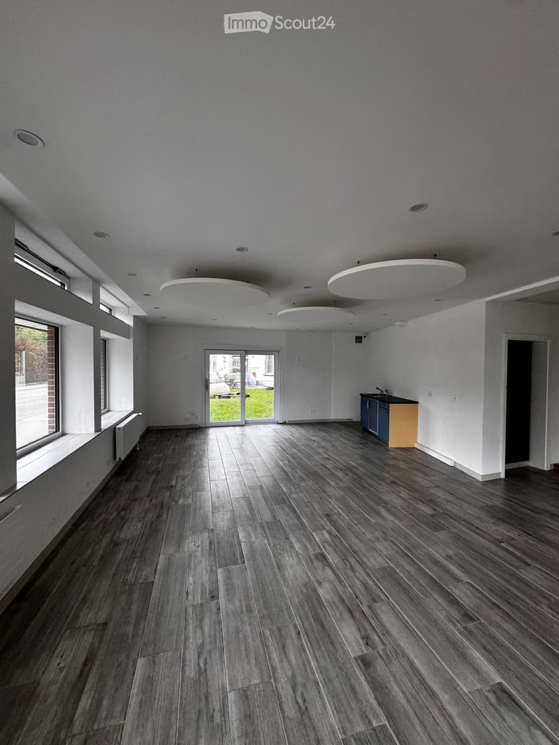 Empty room with wooden flooring, white walls, large windows, and a small kitchen area with cabinets and a sink.