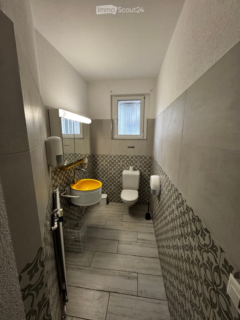 Small bathroom with a toilet, sink, mirror, and decorative patterned tiles. Features a window, light fixture, and a small trash bin.