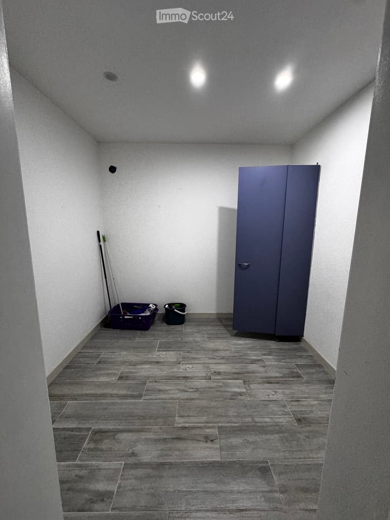 Empty room with grey tiled floor, white walls, and a grey cabinet. Cleaning supplies and a bucket are placed against the wall.