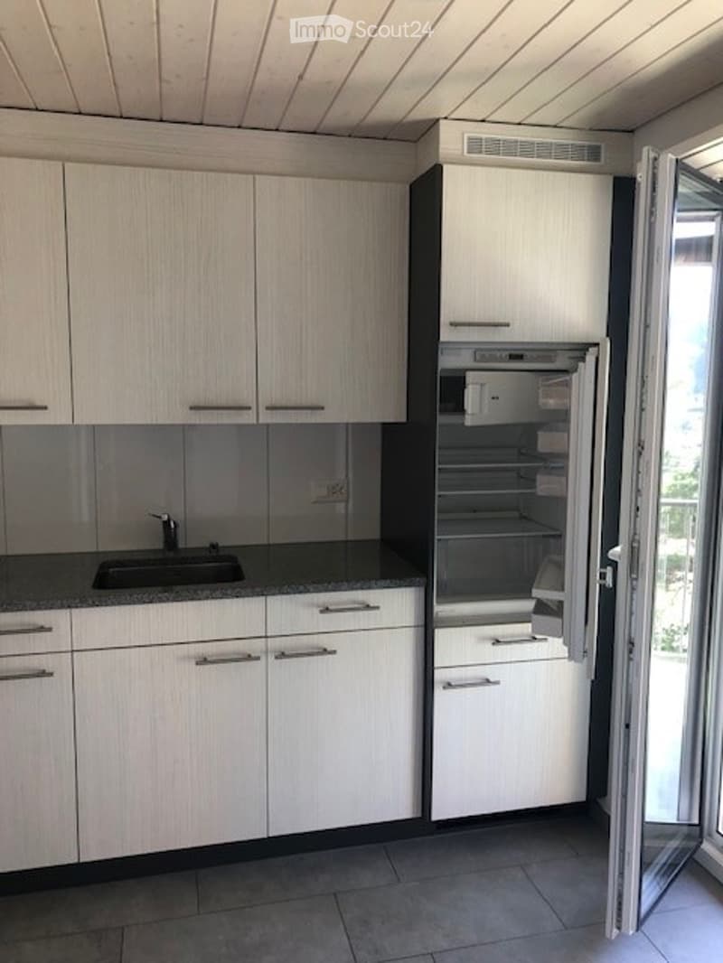 Modern kitchen with white cabinets, black countertop, stainless steel sink, and an open refrigerator. Large window and sliding glass door leading to a balcony.