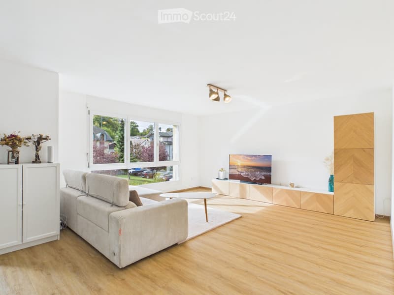Bright living room with large windows, light wooden flooring, grey sectional sofa, coffee table, TV on a wooden stand, and decorative vases.