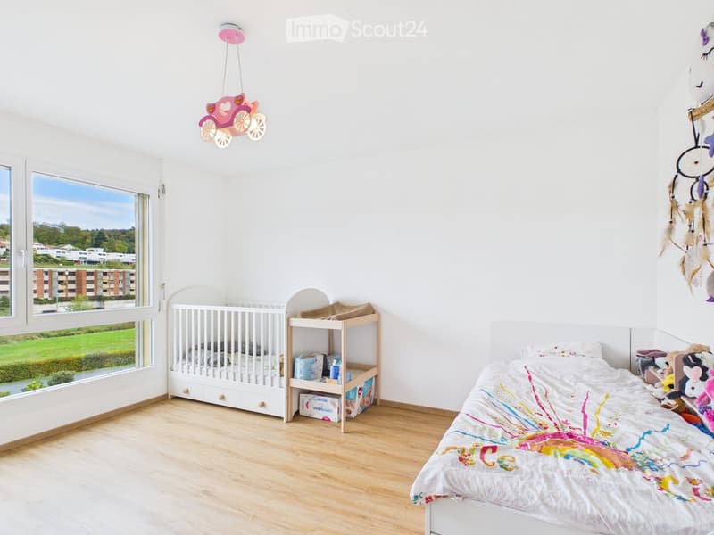 Bright, child-friendly bedroom with a large window offering a view of greenery, a single bed with a colorful duvet, a crib with a changing table, and decorative elements like a dreamcatcher and a whimsical pink car-shaped pendant light.